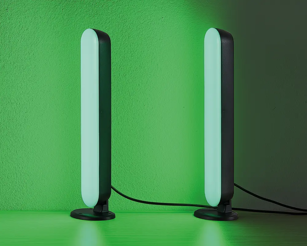 Et par slanke, lodrette LED-lysstænger med afrundede kanter, der udsender et blødt grønt skær. Hver lysstang er monteret på en lille, rund fod og forbundet med en synlig sort strømkabel. Lysene står på en flad overflade op ad en væg og skaber en stemningsfuld belysning.