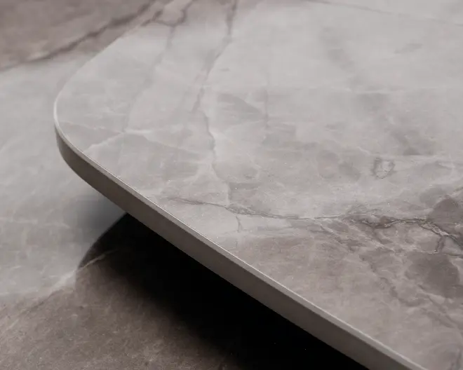 Nærbillede af en bordplade med en glat, poleret overflade i lysegrå marmorlook med diskrete hvide og mørkere grå årer. Kanten er afrundet, hvilket giver et moderne og elegant udtryk. Materialet fremstår solidt og stilfuldt, velegnet til moderne indretning.