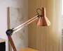 Justerbar skrivebordslampe med en metallisk kobberfinish, konisk lampeskærm og ledarm med synlige fjedre for fleksibel placering. Lampen har et slankt, moderne design og en glat, reflekterende overflade.