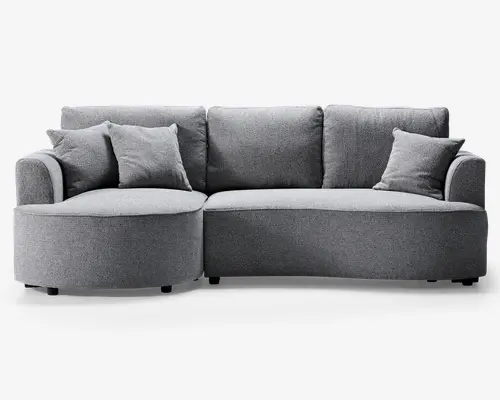 Moderne grå sofa med polstret betræk og et unikt buet design. Sofaen har en chaiselong på venstre side, tre store ryghynder og tre matchende pyntepuder for ekstra komfort. Stoffet ser blødt og tekstureret ud, og sofaen står på lave, næsten usynlige ben, hvilket giver et stilrent og moderne udtryk.