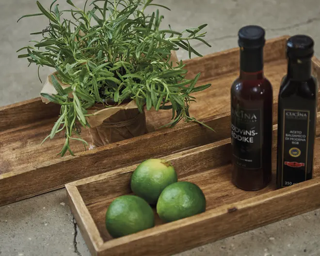 Rektangulære træbakker med naturlig finish og forhøjede kanter. Den ene bakke indeholder en urteplante i potte indpakket i brunt papir, mens den anden bakke har tre limefrugter og to flasker – én med en mørkerød væske og én med en mørk væske, begge med sorte låg og etiketter. Bakkerne har et rustikt, håndlavet udseende og står på en sten- eller betonoverflade.