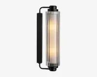Væghængt lampe med cylindrisk glasskærm og lodrette rillede detaljer, der udsender et varmt lys. Lampen har en mat sort metalramme og endestykker, hvilket giver et moderne og industrielt udtryk. Designet er slankt og passer godt til nutidige indretninger.