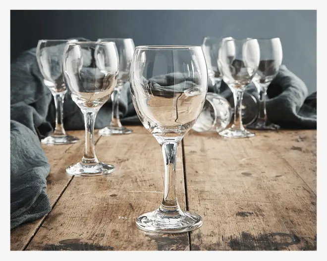 Sæt af klare glasbægre med klassisk rund skål og solid stilk, arrangeret på et rustikt træbord. Glassene har en glat, poleret overflade og en let udadbuet kant, velegnet til servering af vin eller andre drikkevarer. Baggrunden inkluderer et blødt, mørkt stof for en elegant præsentation.