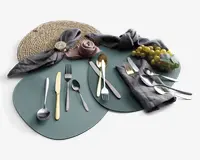 Et sæt bestik med både guldfarvet og sølvfarvet finish, herunder gafler, knive og skeer arrangeret på grønne dækkeservietter. Borddækningen inkluderer grå stofservietter, én rullet som en rose og en anden med en gylden servietring, samt en flettet dækkeserviet og en klase grønne druer som dekoration. Bestikket har et moderne, slankt design med glatte håndtag.