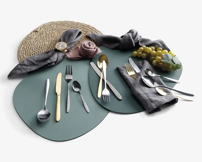 Et sæt bestik med både guldfarvet og sølvfarvet finish, herunder gafler, knive og skeer arrangeret på grønne dækkeservietter. Borddækningen inkluderer grå stofservietter, én rullet som en rose og en anden med en gylden servietring, samt en flettet dækkeserviet og en klase grønne druer som dekoration. Bestikket har et moderne, slankt design med glatte håndtag.