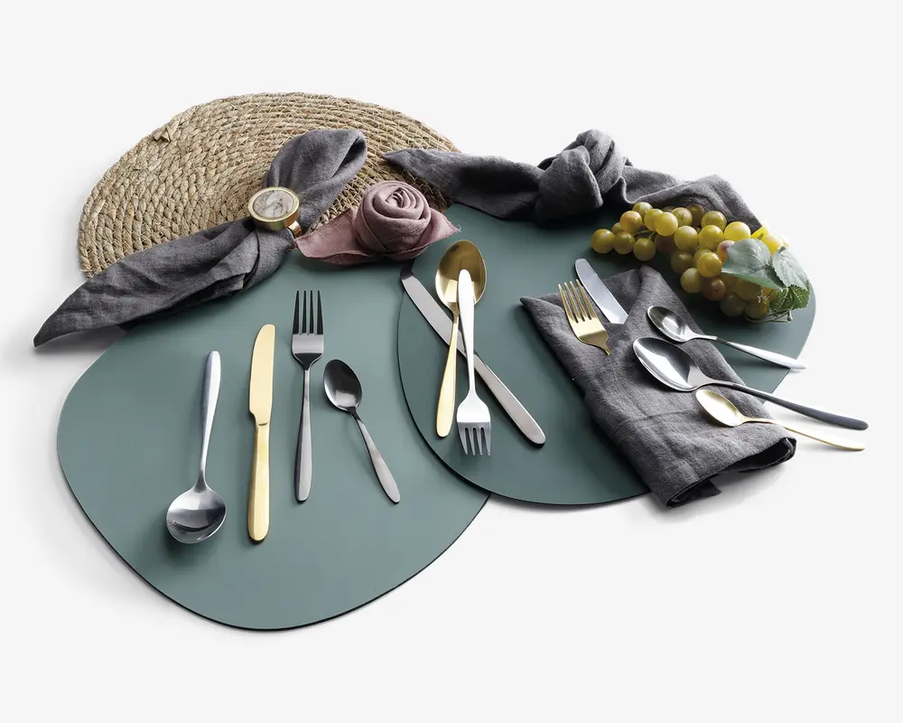 Et sæt bestik med både guldfarvet og sølvfarvet finish, herunder gafler, knive og skeer arrangeret på grønne dækkeservietter. Borddækningen inkluderer grå stofservietter, én rullet som en rose og en anden med en gylden servietring, samt en flettet dækkeserviet og en klase grønne druer som dekoration. Bestikket har et moderne, slankt design med glatte håndtag.