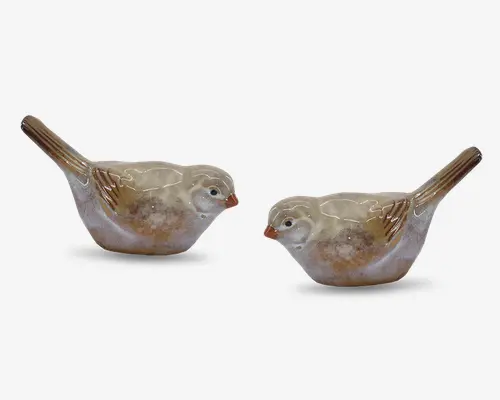 Et sæt af to fugleformede beholdere i keramik med blank overflade. Hver figur er malet i naturlige nuancer af beige, brun og hvid, med detaljerede fjer på vinger og hale. Fuglene har små, runde kroppe, opretstående haler og diskrete ansigtstræk, herunder små næb og malede øjne. De åbne toppe indikerer, at de kan bruges til opbevaring af småting eller som dekorative elementer.