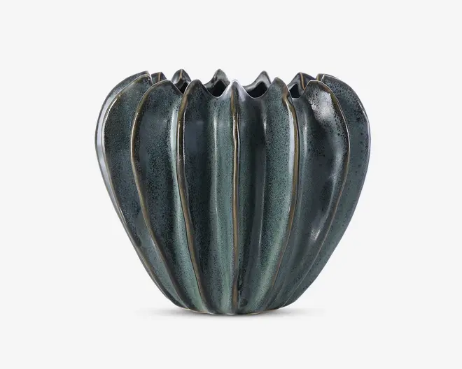 En dekorativ keramisk beholder med blank, mørkegrøn glasur og lodrette riller, der giver et bølget, blomsterlignende udseende. Kanten er ujævn og bølgende, hvilket tilføjer et unikt, organisk præg. Overfladen har diskrete farvevariationer, som giver et håndlavet udtryk.