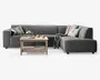 En moderne L-formet hjørnesofa betrukket med mørkegråt stof, med brede armlæn og lav ryg. Sofaen er pyntet med flere dekorative puder i forskellige mønstre og farver samt et hyggeligt tæppe lagt over den ene side. Foran sofaen står et sofabord i lyst træ med en underhylde, hvor der ligger en stak magasiner. Bordpladen har en blomsterdekoration, to krus og en lille tallerken, hvilket skaber en indbydende og komfortabel stueatmosfære.