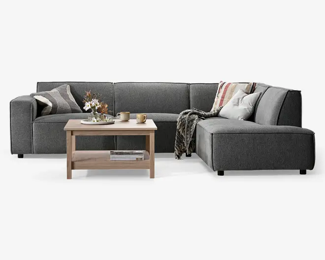 En moderne L-formet hjørnesofa betrukket med mørkegråt stof, med brede armlæn og lav ryg. Sofaen er pyntet med flere dekorative puder i forskellige mønstre og farver samt et hyggeligt tæppe lagt over den ene side. Foran sofaen står et sofabord i lyst træ med en underhylde, hvor der ligger en stak magasiner. Bordpladen har en blomsterdekoration, to krus og en lille tallerken, hvilket skaber en indbydende og komfortabel stueatmosfære.