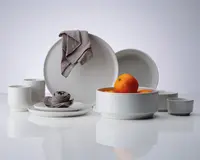 Et sæt minimalistisk, mat hvidt keramikservice bestående af store og små tallerkener, skåle og krus. Delene har glatte, rene linjer og et moderne design. Nogle af skålene er fyldt med appelsiner, og en grå stofserviet er draperet over en tallerken og rullet på en anden, hvilket giver et elegant udtryk til opstillingen.