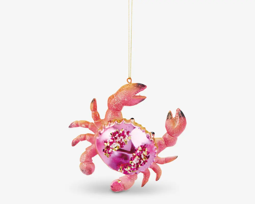 Et dekorativt ornament formet som en krabbe med en blank lyserød krop, guldfarvede detaljer og glimmer. Ornamentet er udsmykket med små blomsterdesigns og glitrende detaljer på skjoldet, og det hænger i en gylden snor. Krabbens ben og kløer er teksturerede og dækket af fint glimmer for en skinnende effekt.