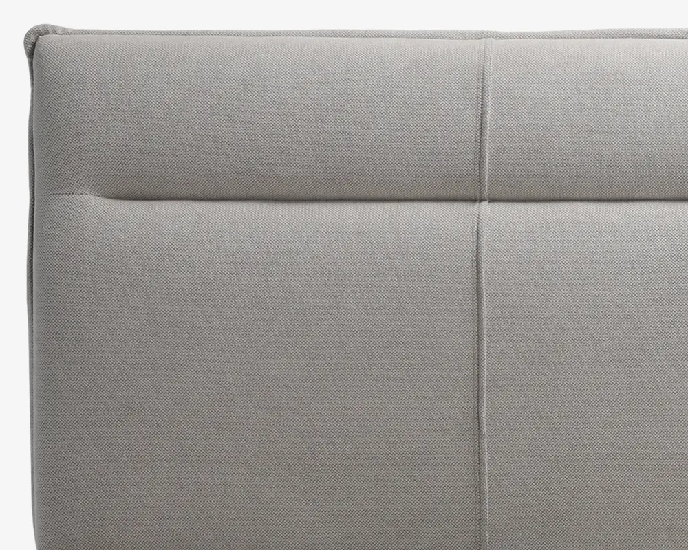 Lys grå polstret overflade med blødt, tekstureret stof. Designet har rene, lige syninger, der deler overfladen op i rektangulære sektioner og giver et moderne og minimalistisk udtryk. Polstringen ser tyk og komfortabel ud med diskrete syningsdetaljer for ekstra stil.