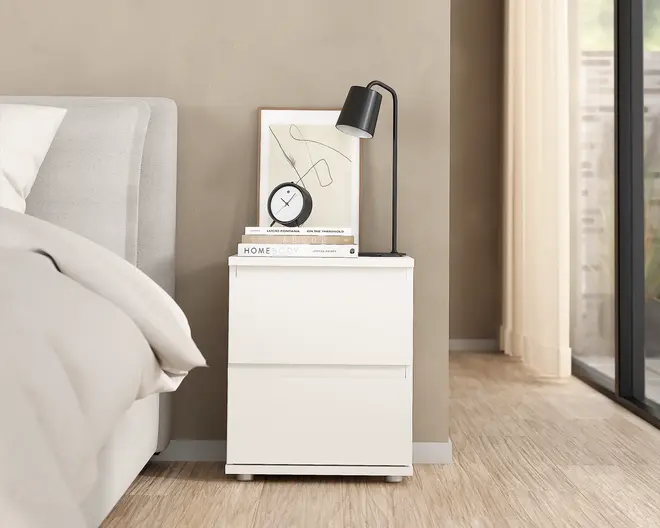 Et moderne, minimalistisk natbord med en glat, mat hvid overflade og to skuffer uden greb. Bordet har en flad top, der er dekoreret med en sort bordlampe, et lille rundt ur, et indrammet abstrakt line-art billede og en stak bøger. Designet er enkelt og stilrent, velegnet til nutidige indretninger.