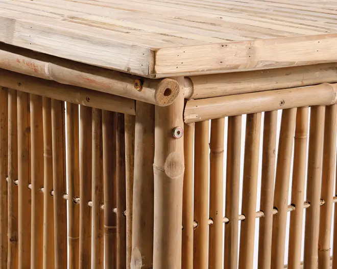 Nærbillede af et møbel lavet af naturlig bambus. Konstruktionen har lodrette bambusstænger på siderne, med vandrette bambuspinde, der forstærker kanter og hjørner. Toppen er flad og består af tæt placerede bambuslameller. Bambussen har en glat, lys finish, der fremhæver dens naturlige åretegning og farvevariationer. Synlige skruer og samlinger viser det håndlavede arbejde.