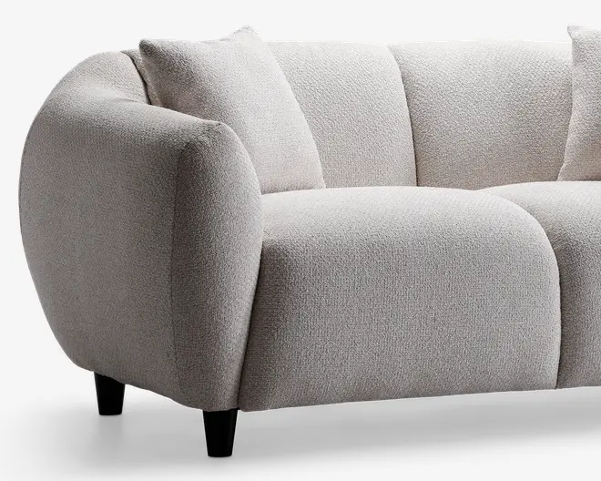 Buet armlæn og ryglæn med blødt, tekstureret stof i en lys beige farve. Sæde- og ryghynderne er tykke og generøst polstrede for ekstra komfort. Betrækket har en blød, boucle-lignende overflade, og sofaen står på korte, koniske sorte ben. Designet er moderne med afrundede kanter og et hyggeligt, indbydende udtryk.