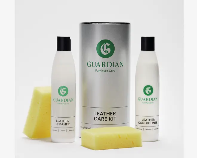 Billedet viser et læderplejesæt bestående af to høje flasker med sorte låg, mærket 'Leather Cleaner' og 'Leather Conditioner'. Begge flasker er hvide med grøn og sort tekst. Sættet indeholder også en stor, cylindrisk sølvbeholder med mærket 'Guardian Furniture Care' samt to gule, rektangulære svampe. Emballagen er enkel og moderne med tydelig mærkning af hvert produkt.
