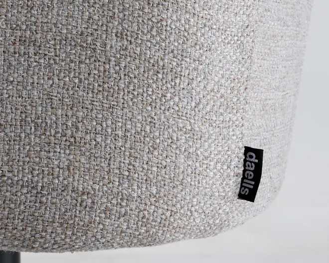 Nærbillede af et tekstureret, lysebeige vævet stof med en diskret blanding af creme- og lysebrune tråde. Stoffet fremstår tæt vævet og slidstærkt. Et lille sort stofmærke med mærkenavnet 'daells' i hvide små bogstaver er syet ind i sømmen.