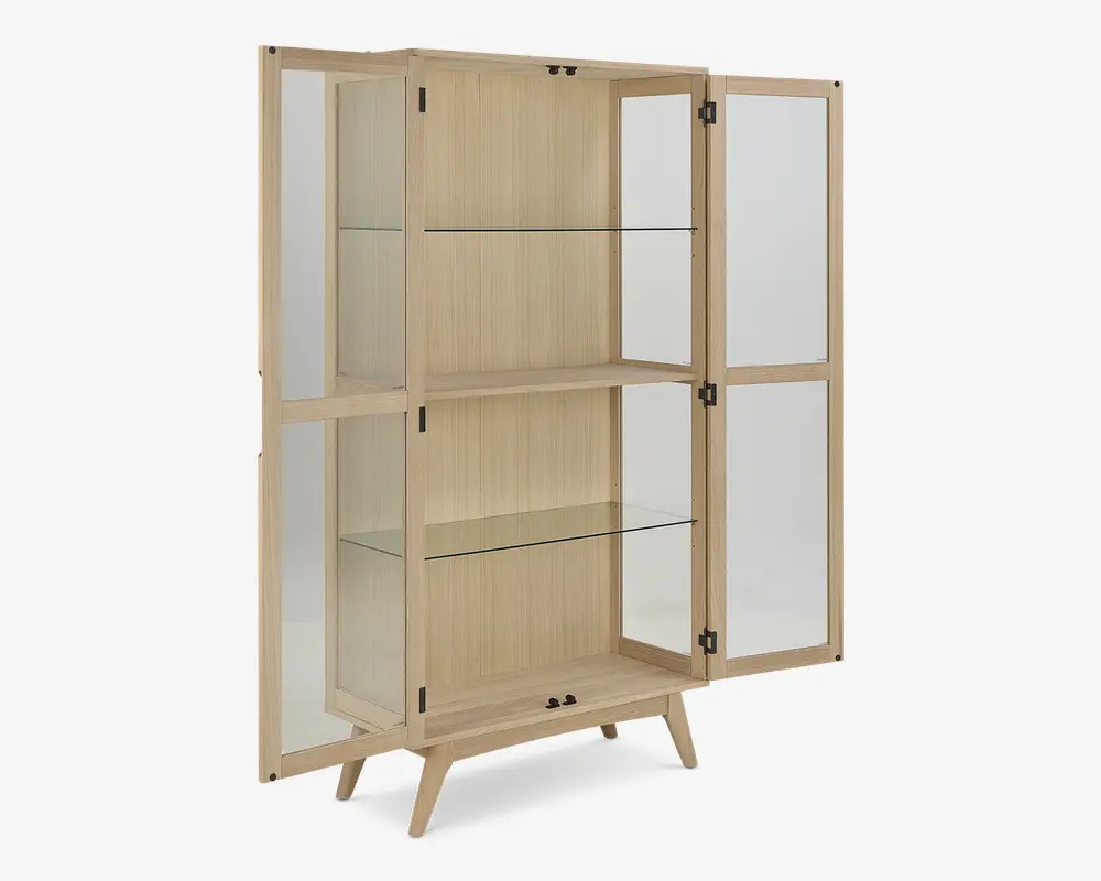 Høj vitrineskab med lys træramme, to glaslåger og glaspaneler på siderne. Indvendigt er der to klare glashylder til opbevaring eller udstilling, og skabet står på fire skråtstillede ben for et moderne udtryk. Lågerne har små runde greb, og designet er minimalistisk og elegant.