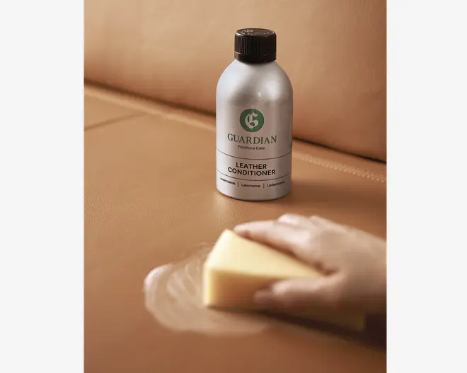 En sølvfarvet flaske med sort låg, mærket 'Guardian Furniture Care Leather Conditioner', står på en lysebrun læderoverflade. I forgrunden ses en hånd, der bruger en gul svamp til at påføre læderplejemidlet i cirkulære bevægelser, hvilket giver et let blankt område på læderet. Billedet fremhæver produktets anvendelse til pleje og vedligeholdelse af læder.