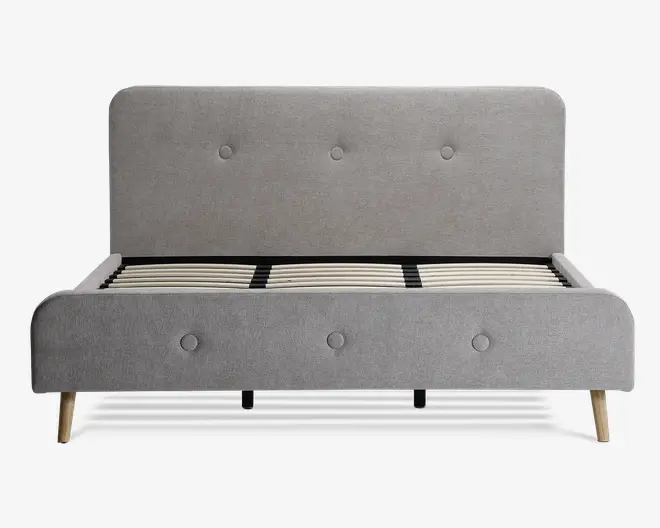 Moderne sengegavl med lysegråt polstret stof, der har en høj, afrundet hovedgavl og matchende fodgavl, begge med knapdetaljer. Rammen har trælameller til madrasstøtte og står på skråtstillede lyse træben for et moderne udtryk.