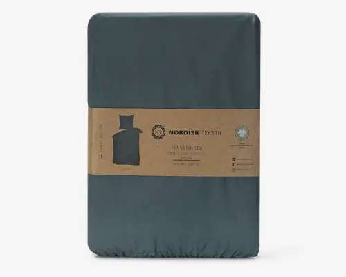 Mørk teal sengetøjssæt fremstillet af økologisk bomuld, pakket i en rektangulær form med et brunt papirbånd omkring midten. Båndet viser Nordisk Tekstil-logoet, produktinformation og en lille illustration af sengetøjet. Emballagen fremhæver, at materialet er GOTS-certificeret økologisk bomuld og inkluderer størrelsen 140x200/60x63 cm.