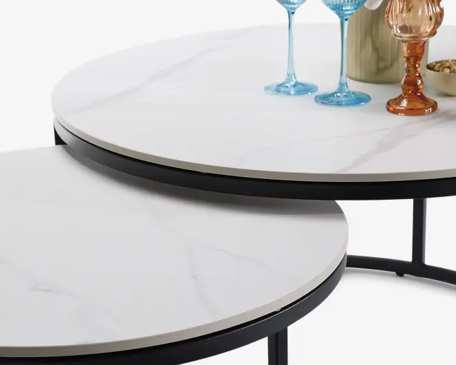 Nærbillede af to runde bordplader med hvid marmorlignende overflade og diskrete grå årer. Kanterne er let skrå og indrammet af en tynd sort kant. Bordene er placeret, så det ene delvist kan skubbes ind under det andet. På det øverste bord står farvet glas og en lille skål, hvilket giver et elegant udtryk.
