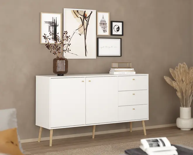 Et moderne skænk med mat hvid finish, der har to skabslåger til venstre og tre skuffer til højre, alle med små gyldne greb. Skænken står på fire skrå ben i lyst træ, hvilket giver et retro-inspireret udtryk. Toppen er dekoreret med en brun glasvase med tørrede grene samt en stak bøger med et gyldent dekorativt fad. Baggrunden er en taupefarvet væg med en billedvæg af indrammede abstrakte og arkitektoniske kunsttryk.