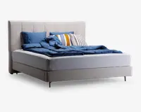 Moderne seng med et lysebeige polstret hovedgærde og base, der har et rent og minimalistisk design. Sengen er redt med blåt sengetøj, herunder dyne og puder, og pyntet med dekorative puder i blå, hvide og gule stribede mønstre. Sengen står på slanke metalben, hvilket giver et moderne udtryk.