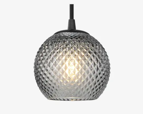 Rund pendellampe med en tekstureret glasskærm i diamantmønster. Glasset er klart, hvilket gør det muligt for den varme glød fra pæren indeni at skinne igennem. Lampen hænger fra en sort ledning og har en metalhætte øverst. Designet giver et moderne og elegant udtryk, og det mønstrede glas skaber en glitrende effekt, når lampen er tændt.