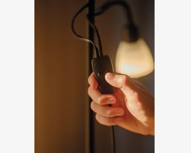 Nærbillede af en hånd, der holder en sort ledningsafbryder med trykknap, som sidder på en ledning. I baggrunden ses en lampe, der lyser blødt. Afbryderen har et enkelt design, der gør den nem at betjene.