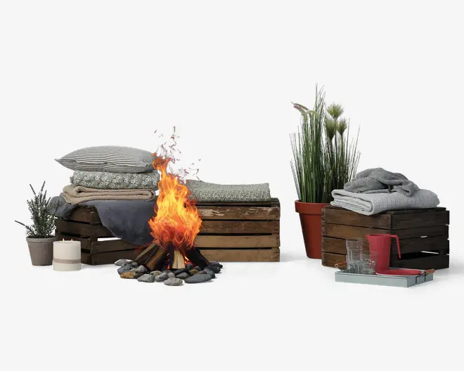 Et hyggeligt udendørsarrangement med sammenfoldet sengetøj i både mønstrede og ensfarvede lagner og tæpper, stablet på rustikke trækasser. I midten er der et lille bål omgivet af sten. Scenen er pyntet med potteplanter, et hvidt stearinlys og en bakke med to klare glas og et rødt krus, hvilket skaber en varm og indbydende stemning.