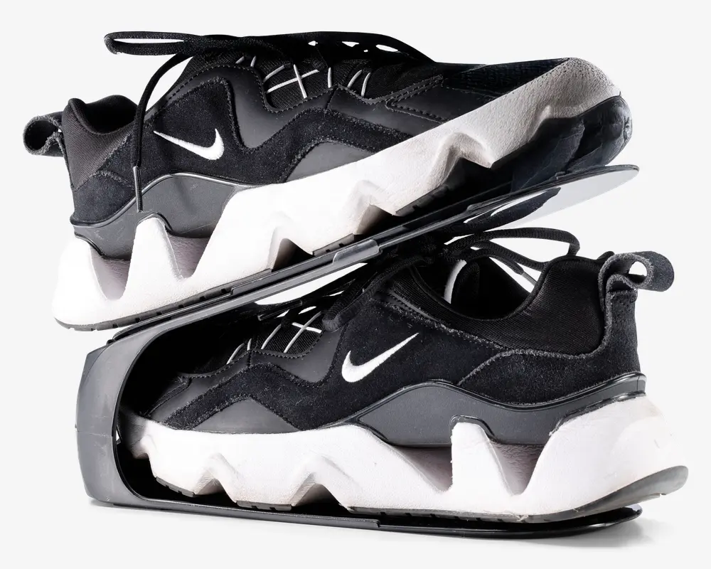 Par sorte og hvide sportssko med en kraftig, zigzag-mønstret hvid sål og sort overdel med ruskinds- og meshpaneler. Skoene har et lille hvidt swoosh-logo på siden, sorte snørebånd og en strop bagpå hælen. Skoene er vist stablet sål mod sål, hvilket fremhæver den tykke, skulpturelle mellemsål og den teksturerede ydersål.