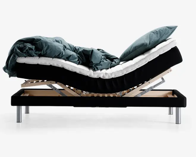 Sidebillede af en justerbar seng med en opdelt madras, vist i en hævet position ved både hoved- og fodende. Sengen har en solid træramme med metalben og en synlig lamelbund for støtte. Madrassen er dækket af en tyk, blød dyne og puder i mørkegrøn farve, hvilket giver et hyggeligt og indbydende udtryk.