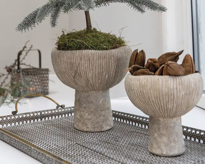To teksturerede skåle på fod med riflede sider og stenlook, den ene fyldt med mos og et lille træ og den anden med dekorative frøkapsler, placeret på en metalbakke.