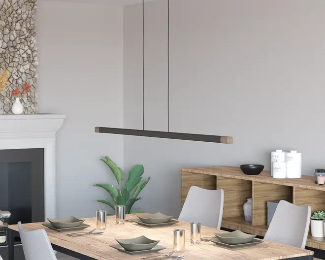 Et moderne, minimalistisk pendellampe med en lang, slank rektangulær form, ophængt fra loftet med to tynde sorte ledninger. Lampen har en mat sort finish med naturlige trædetaljer i enderne, hvilket giver et stilrent og nutidigt udtryk. Den er designet til at give jævn belysning over spisebordet nedenunder.