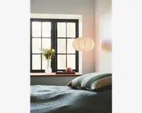 Et blødt oplyst soveværelse med en stor, rund, hvid papirlampe i lanterne-stil, der hænger fra loftet. Lampen har en rillet struktur og udsender et varmt, diffust lys. Under lampen er der et vindue med sorte rammer og en vindueskarm af træ. På karmen står en hvid keramisk vase med en buket friske blomster, en lille lyserød pyntegenstand og en lukket bog. Sengen i forgrunden er redt med et tekstureret, lyseblågrønt tæppe og har puder med grønne og hvide striber.