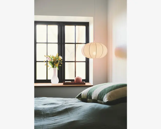 Et blødt oplyst soveværelse med en stor, rund, hvid papirlampe i lanterne-stil, der hænger fra loftet. Lampen har en rillet struktur og udsender et varmt, diffust lys. Under lampen er der et vindue med sorte rammer og en vindueskarm af træ. På karmen står en hvid keramisk vase med en buket friske blomster, en lille lyserød pyntegenstand og en lukket bog. Sengen i forgrunden er redt med et tekstureret, lyseblågrønt tæppe og har puder med grønne og hvide striber.