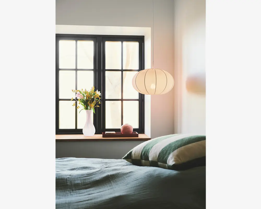 Et blødt oplyst soveværelse med en stor, rund, hvid papirlampe i lanterne-stil, der hænger fra loftet. Lampen har en rillet struktur og udsender et varmt, diffust lys. Under lampen er der et vindue med sorte rammer og en vindueskarm af træ. På karmen står en hvid keramisk vase med en buket friske blomster, en lille lyserød pyntegenstand og en lukket bog. Sengen i forgrunden er redt med et tekstureret, lyseblågrønt tæppe og har puder med grønne og hvide striber.