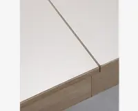 Nærbillede af et bordhjørne med en lys træramme og en glat, mat hvid bordplade. Bordpladen har en synlig samling, hvilket tyder på, at den kan være udtrækkelig eller modulopbygget. Træets årer er diskrete, og designet er minimalistisk og moderne.