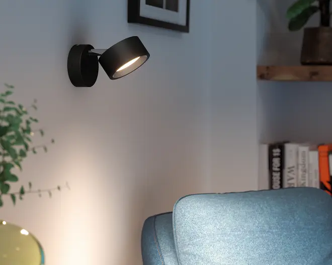 Sort vægmonteret justerbar spotlight med rund base og vinklet cylindrisk lampeskærm, vist tændt med varmt lys.