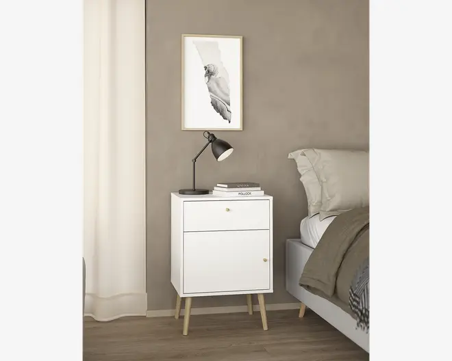 Et moderne, hvidt natbord med rene linjer, der har en enkelt skuffe og en skabslåge, begge med små runde greb. Natbordet står på fire skråtstillede ben i lyst træ. På toppen står en sort skrivebordslampe og en lille stak bøger, og overfladen er glat og ryddelig. Designet er minimalistisk og inspireret af skandinavisk stil.