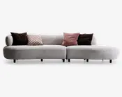 Moderne, lysegrå sofa med lav ryg og afrundede kanter. Sofaen har en chaiselong-del til højre og står på korte, sorte ben. Den er pyntet med fem dekorative puder: to ensfarvede mørkebrune, én lyserød med geometrisk mønster, én ensfarvet lyserød og én med cirkelmønster i beige og brune nuancer. Betrækket ser blødt og behageligt ud, hvilket giver en komfortabel siddeoplevelse.