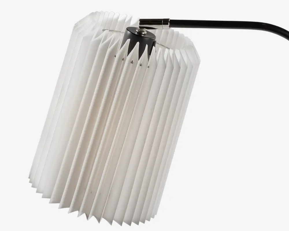 Et cylindrisk lampeskærm med plisseret, harmonika-lignende design lavet af hvidt materiale. Skærmen er monteret på et sort metalbeslag i toppen, hvilket giver et moderne og tekstureret udtryk. Plisseringerne skaber et geometrisk mønster og giver en blød lysspredning.