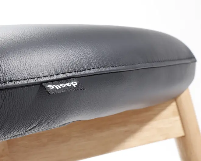 Nærbillede af en sædepude betrukket med sort kunstlæder med synlig struktur. Puden har en pænt syet kant og en lille sort stofmærke med hvid tekst. Sædet hviler på en lystræramme, hvor en del af benet og støtte er synlig.