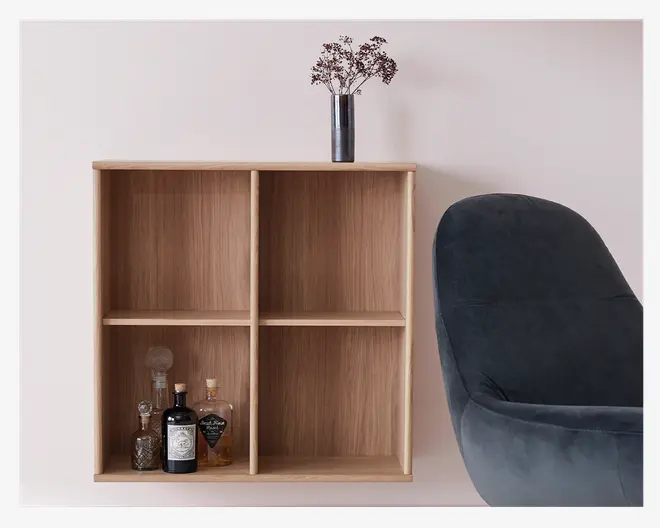 En vægmonteret reol i lyst træ med fire kvadratiske rum. Hylderne har en glat overflade med naturlig træstruktur. Nederste venstre rum indeholder flere glasflasker, herunder en karaffel og spiritusflasker. Ovenpå reolen står en slank, mørk vase med tørrede blomster. Til højre ses en del af en blød, mørkegrå polstret stol.