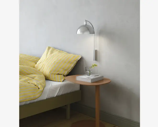 Et sengebord med en lille rund træbordplade i lys naturfarve. På bordet står et klart glas vand, en stak på to hvide hardback-bøger og en lille glasvase med en enkelt grøn kvist. Sengen ved siden af bordet har sengetøj med gule og hvide striber, herunder en pude og dyne. Over bordet er der monteret en moderne væglampe med buet metalskærm, som giver fokuseret belysning.