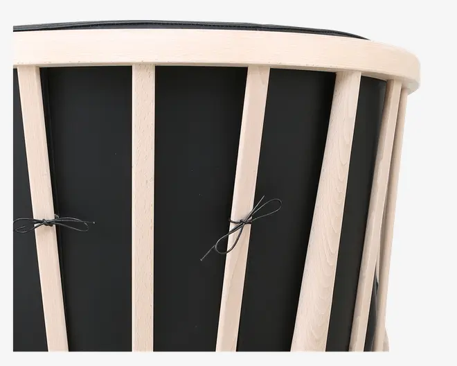 Nærbillede af et produkt med lodrette lyse trælameller, der er jævnt fordelt omkring en sort cylindrisk overflade. Lamellerne er fastgjort til en afrundet træramme øverst, og sorte snore er bundet i sløjfer mellem nogle af lamellerne, hvilket antyder et dekorativt eller funktionelt element. Designet er moderne og minimalistisk.