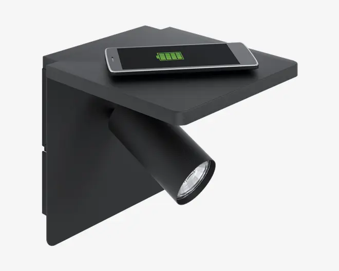 Væghængt armatur i mat sort med en rektangulær hylde øverst og en indbygget spotlampe, der peger skråt nedad. Hylden har en smartphone liggende ovenpå, som trådløst oplades, hvilket ses på batteriikonet på telefonens skærm. Designet er moderne og minimalistisk, velegnet til brug ved seng eller læseplads.