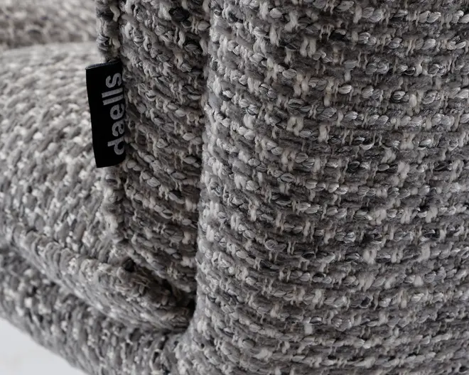 Nærbillede af et tekstureret, vævet stof i grå og offwhite nuancer med en tyk, blød og let grov vævning. Et lille sort stofmærke med mærket 'daells' i hvid tekst er syet ind i sømmen. Stoffet ser blødt og behageligt ud, velegnet til polstring.